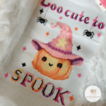 Kreuzstich Vorlagen Kürbis mit rosafarbenen Hexenhut umrandet von bunten Funkelsternen. Über dem Kürbis steht der Schriftzug Too cute to in rosa und schwarz mit zwei sich abseilenden Spinnen. Unter dem Kürbis steht in Großbuchstaben SPOOK in bunten Farben und schwarzen Schatten. Das Bild ist seitlich aufgenommen und dekoriert mit künstlichen Spinnweben und etwas Garn