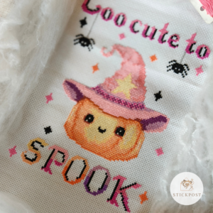 Kreuzstich Vorlagen Kürbis mit rosafarbenen Hexenhut umrandet von bunten Funkelsternen. Über dem Kürbis steht der Schriftzug Too cute to in rosa und schwarz mit zwei sich abseilenden Spinnen. Unter dem Kürbis steht in Großbuchstaben SPOOK in bunten Farben und schwarzen Schatten. Das Bild ist seitlich aufgenommen und dekoriert mit künstlichen Spinnweben und etwas Garn