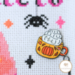 Cozy Pumpkin Mug Nadelmagnet als Stickzubehör zum Kreuzstich sticken. Ein Becher mit herbstlichen Kürbismotiv und Sahnehaube. Angewendet auf einem Stickbild. Zeigt wie die Magnethalterung die Sticknadel hält.