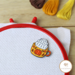 Cozy Pumpkin Mug Nadelmagnet als Stickzubehör zum Kreuzstich sticken. Ein Becher mit herbstlichen Kürbismotiv und Sahnehaube. Magnethalterung auf Stoff im Stickrahmen gezeigt.