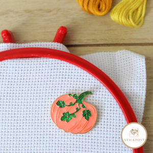 Herbstkürbis Nadelmagnet. orangefarbener Kürbis mit grünen Blätterranken. Magnethalterung auf Stoff im Stickrahmen gezeigt.