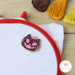 Pumpkin Kitty Nadelmagnet. Lila und rosafarbene Kürbiskatze im Jack-o-lantern Stil. Magnethalterung auf Stoff im Stickrahmen gezeigt.